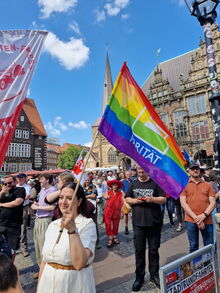 Junge Frau mit bunter Flagge und Parität-Logo auf dem Bremer Marktplatz , im Hintergrund viele Menschen