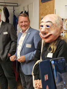 Eine Person hat einen großen Maskenkopf auf, daneben ein lachender Mann