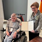 Ein älterer Mann im Rollstuhl, hält auf dem Schoß eine Schachtel mit einem Bundesverdienstkreuz, daneben eine Frau mit Urkunde lächelnd.