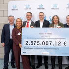 7 Personen in Anzügen halten einen großen Scheck über 2,757 Mio. Euro in der Hand