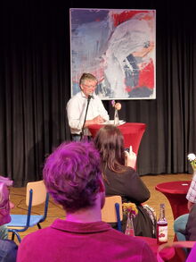 Mann um die 65 steht an einem runden Tisch und spricht in ein Mikro, im Vordergrund sitzen Gäste an kleinen Tischen mit Blumen und Getränekn