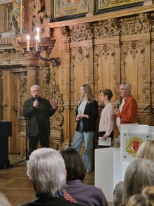 Ein älterer Mann in schwarz mit Mikro, daneben 3 Personen, im Hintergrund Holzvertäfelung.