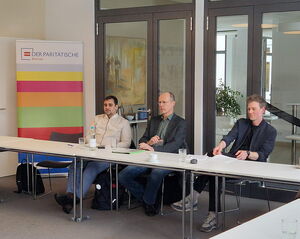 3 Männer sitzen an Tischen eines Seminarraums, im Hintergrund ein Roll-up des Paritätischen Bremen