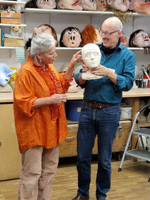 Eine Frau und ein Mann halten eine Gipsmaske in der Hand, im Hintergrund im Regal viele Maskengesichter