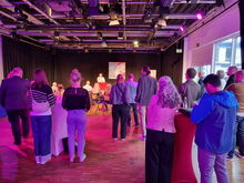 Im Vordergrund stehen Gäste an Stehtischen, dahinter sitzen Menschen an Tischen, im Hintergrund ein Mann am Mikro