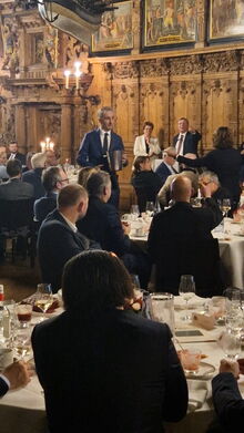 Männer in schwazen Anzügen sitzen an festliche Tischen, ein Mann geht mit einem Eimmer herum