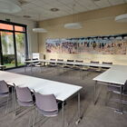 Heller Seminarraum mit weißen Tischen im Carree gestellt, links großes Fenster mit Grün, Gemälde aus 4 Bildern an der Wand