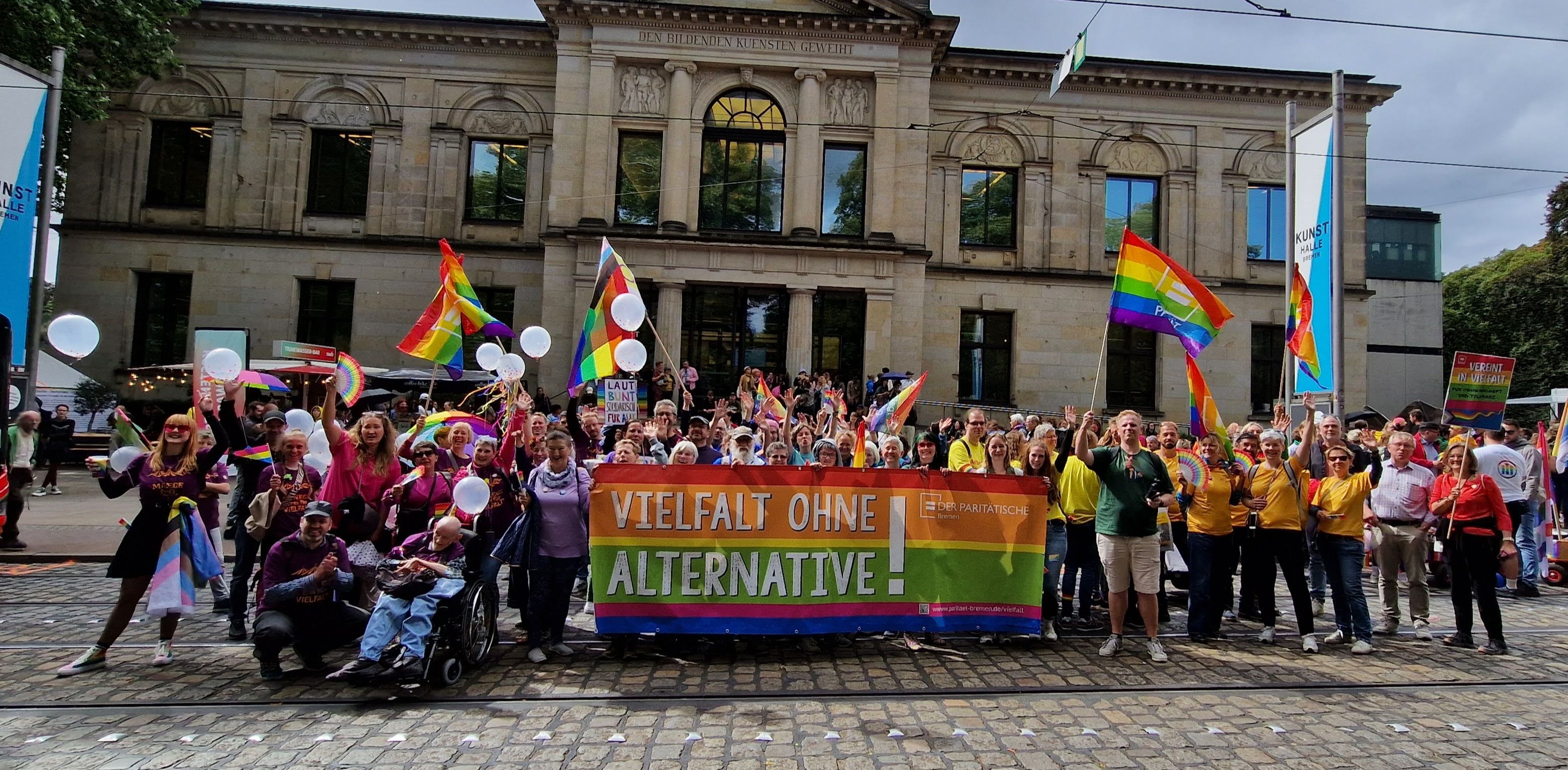 großes buntes Banner "Vielfalt ohne Alternative" und Logo des Paritätischen, dahinter und daneben viele Menschen mit bunten Flaggen bei einer Demo (CSD), stehend vor der Kunsthalle