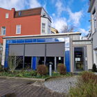 Gebäude mit blauer Fassade und Sonnenschutz, im Hintergrund rotes Gebäude, vorne Bepflanzung und Plastersteine