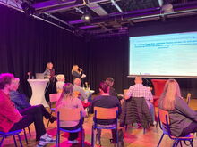 mehrere Menschen sitzen und schauen auf eine Leinwand, links zwei Moderatorinnen. Text auf der Leinwand: Zu welcher Frage würden Sie sich gern fachübergreifend austauschen und vernetzen? mit unlsebaren Antworten darunter