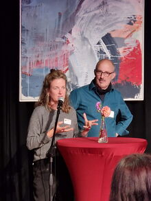 Eine Frau und ein Mann stehen an einem runden Tisch mit Mikro, dahinter ein dunkler Vorhang und ein Gemäde