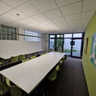 Seminarraum mit großer Tafel aus mehreren Tischen, grüne Stühlen, grüne Wand mit Bildern, links Wand mit Glasbausteinen, Fensterfront mit Terrassentür