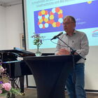 Mann Mitte 60 an einem Stehtisch mit Mikro, im Hintergrund eine Leinwand mit einer Präsentation, ein Piano und Blumen