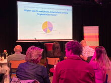 Menschen auf Stühlen blicken auf eine Leinwand mit einem Kuchendiagramm, Titel: Interaktiv: In welchem Arbeitsfeld ist Ihre Organisation tätig?