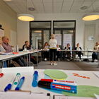 Seminarsituation: im Vordergrund Stifte, Edding, grüne Zettel, im Hintergrund unscharf mehrere sitzende Personen an Tischen, eine Frau stehend