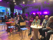 Veranstaltungsraum: Menschen sitzen und stehen an runden Tischen und unterhalten sich