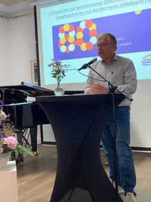 Mann Mitte 60 an einem Stehtisch mit Mikro, im Hintergrund eine Leinwand mit einer Präsentation, ein Piano und Blumen
