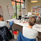 Seminarraum mit weißen Tischen, 3 Menschen sichtbar, viele Stellwände, 2 Flipchart und bunte Zeichnungen und Zettel